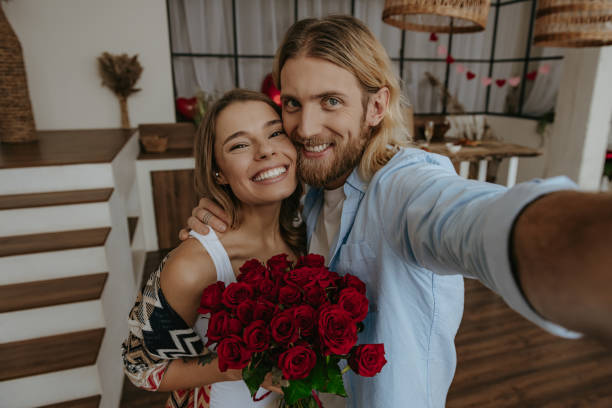 istock Smiling young man making selfie while embracing his beautiful girlfriend holding a bouquet of flowers at home 1930228361