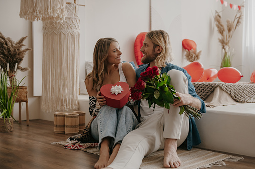 Loving couple embracing and holding roses and gift box while spending Valentines Day at cozy home