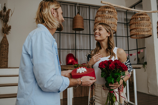 Handsome young man giving a flower bouquet and gift box to his surprised girlfriend standing at home