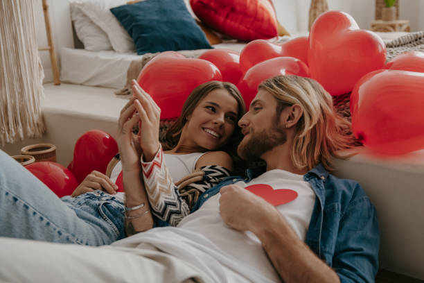 istock Young loving couple holding heart shape Valentines cards while leaning on bed surrounded with red balloons 1930238027