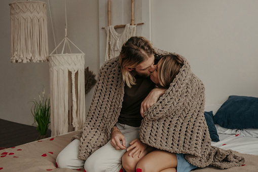 Happy young loving couple covering with a cozy warm blanket and kissing while sitting on bed together