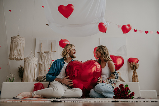 Happy loving couple holding big red heart shape balloon while celebrating Valentines day at cozy home