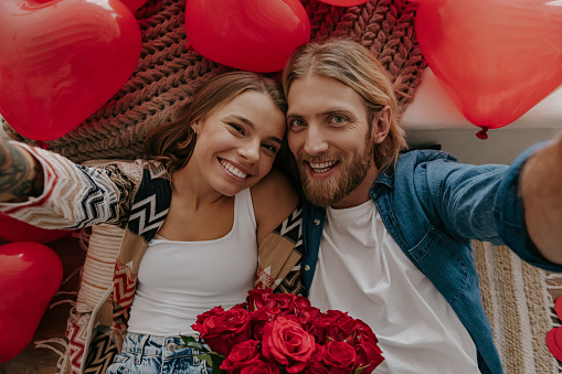 Top view of happy loving couple holding flowers and making selfie with red heart shape balloons around them
