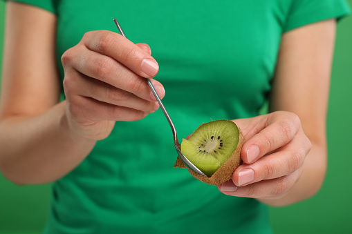 Woman eating kiwi with spoon on green background, closeup