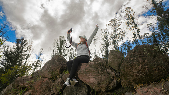 A young woman joyfully poses on a cluster of giant rocks, raising her hands in the air in a carefree and celebratory manner