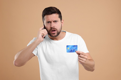 Emotional man with credit card talking on smartphone against beige background. Be careful - fraud