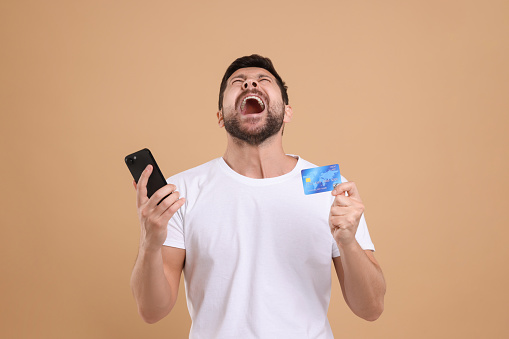 Emotional man with credit card and smartphone on beige background. Be careful - fraud