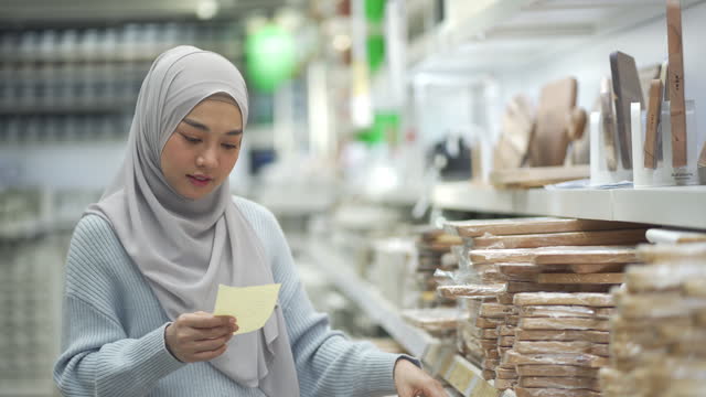 4K Young Muslim Woman Checking A Shopping List On Paper And Choosing Kitchen utensils In A Shopping Mall