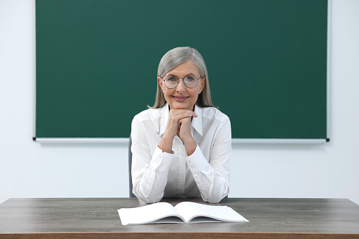 beautiful teacher with glasses sits at a desk with book, pencil notepad apple against blackboard, classroom