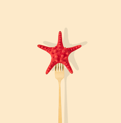 Red starfish pricked on a golden fork on light pastel cream background. Minimal summer concept. Creative trendy sea star with fork idea. Summer aesthetic.