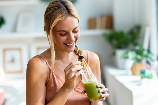 Shot of happy woman drinking a healthy green smoothie standing in the living room at home.