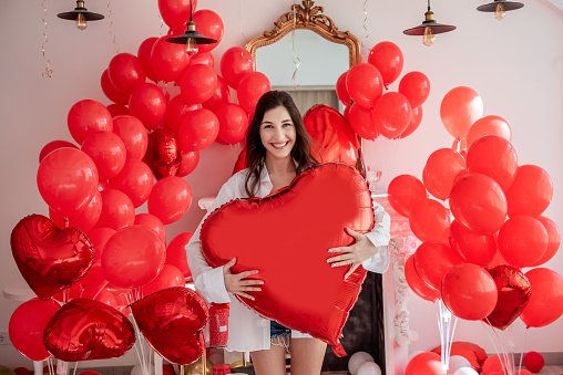 Young woman smiling while standing in room decorated for celebration Valentines Day. Girl holds large red heart shaped balloon. Rose petals scattered on floor along with pillows gift boxes. Mock up