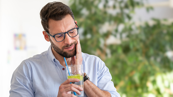 A man drinks a cold drink with ice through a straw and feels severe pain in his teeth due to damage to the tooth enamel. Lie down and hold the place of pain. He removes a straw and ice cubes from a glass of cold drink.