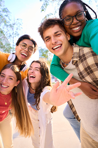 Vertical multiracial group of teenagers taking a selfie looking front camera laughing and having fun piggybacking together outdoor. Young students enjoying their free time with friends in street city