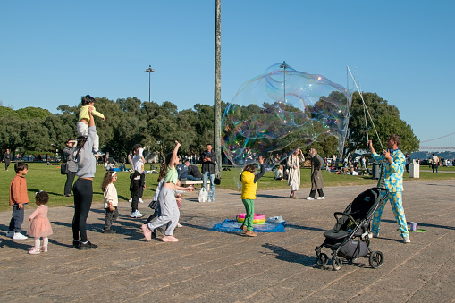 In Lisbon, Portugal, joyful children play with soap bubbles on the city's main square, while happy locals and tourists bask in the warmth of a sunny evening.