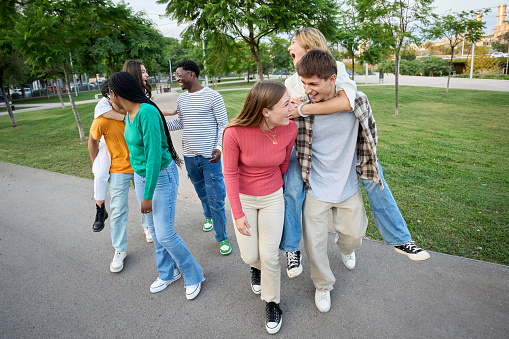 Cheerful multiracial group of people laughing and having fun piggybacking together in an outdoor garden area. Young students enjoying their leisure and free time with friends in the park