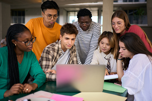 Concentrated young group of university student using laptop in the cafeteria on campus with his classmates working in a project. Academic multiracial people studying together on the faculty building