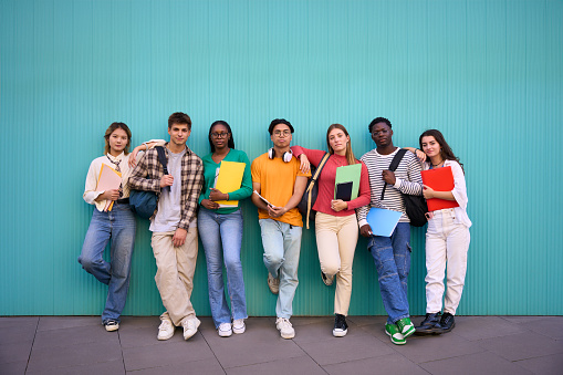 Serious international students posing standing looking at the camera. Young people multicultural friends concentrated and embracing with backpacks and workbooks in a blue wall background
