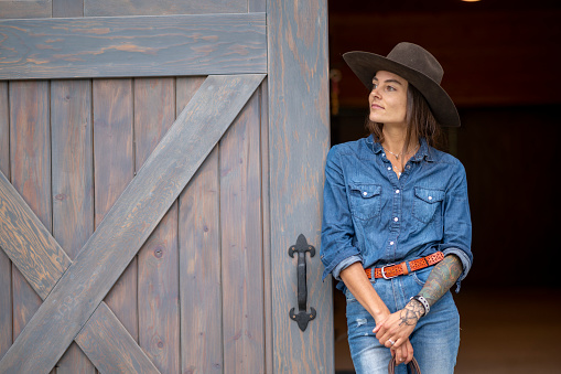 Cowgirl leans against barn door and looks off