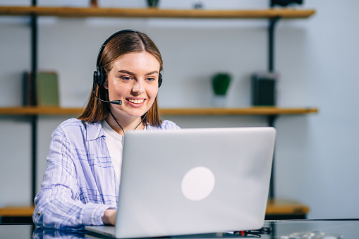 Happy woman with headphones watching webinar on laptop in modern office, smiling young business woman having fun, laughing, talking on video call with client or colleague using wireless internet connection