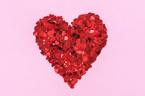 Heart made of shiny red small decorative hearts on a pink background. Flat lay, place for text. Festive minimal composition.