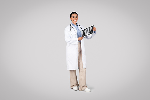 Latin woman doctor in professional attire, holding and examining an X-ray of the lungs, showing careful medical observation and diagnostic process, grey studio background