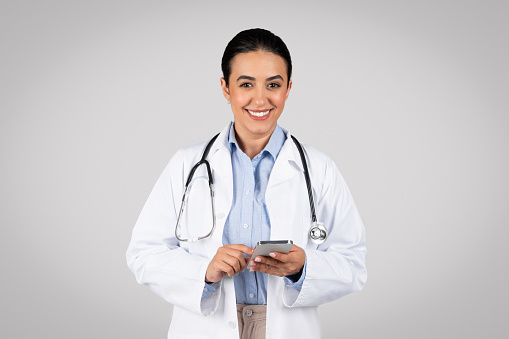 Portrait of friendly latin female therapist in white coat and with stethoscope using cellphone for work and smiling at camera, gray background. Health care and treatment remotely, app for medicine