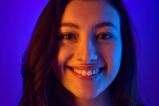 Close up portrait of young attractive woman smiling and looking at camera against gradient blue background in neon light. Concept of positive emotions, fashion and beauty, wellness. Ad
