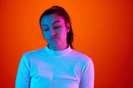 Feel guilty. Young woman wearing white long sleeve in bad mood, looked down against gradient orange-red background in neon light. Concept of mistake, unwell, sadness, frustration, mental health. Ad