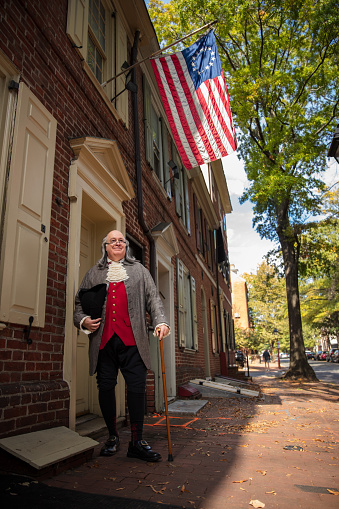A full body shot of an actor impersonating Benjamin Franklin standing under an American flag on a sunny day in Philadelphia. Featuring Brian Patrick Mulligan as Ben Franklin.