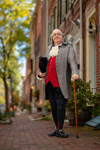 A full body shot of an actor impersonating Benjamin Franklin standing on the sidewalk with his hat and a walking cane on a sunny day in Philadelphia. Featuring Brian Patrick Mulligan as Ben Franklin.