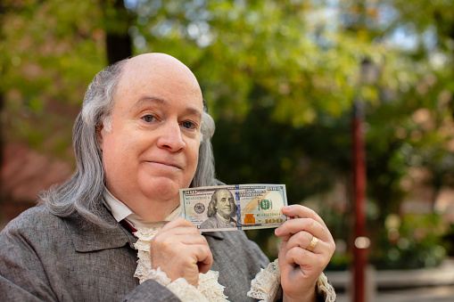 A close up shot of an actor impersonating Benjamin Franklin holding a hundred dollar bill near his face, while out on a sunny day in Philadelphia. Featuring Brian Patrick Mulligan as Ben Franklin.