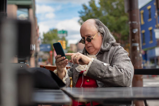 Benjamin Franklin Impersonator Tapping his Smartphone While Sitting in Outdoor Patio of Bar A medium close up shot of an actor impersonating Benjamin Franklin, with earphones plugged in, using his phone while sitting in an outdoor patio in modern-day Philadelphia. Featuring Brian Patrick Mulligan as Ben Franklin. patriotic man stock pictures, royalty-free photos & images