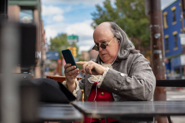 sobowtór benjamina franklina stuka w smartfona, siedząc na zewnętrznym patio baru - benjamin franklin history american culture portrait zdjęcia i obrazy z banku zdjęć