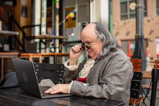 A medium close up shot of an actor impersonating Benjamin Franklin working on a laptop while sitting in an outdoor patio on a sunny day in modern-day Philadelphia. Featuring Brian Patrick Mulligan as Ben Franklin.