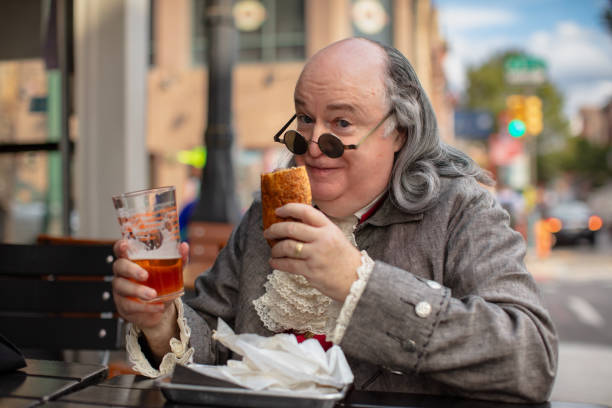 medium close up of benjamin franklin impersonator with philly cheesesteak and glass of beer - benjamin franklin history american culture portrait imagens e fotografias de stock