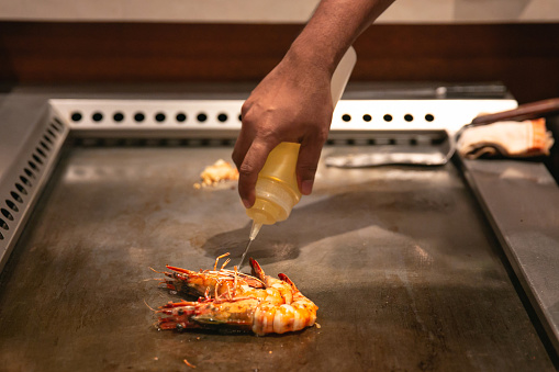 Close-up shot of unrecognizable teppanyaki chef pouring oil on top of a tiger prawn ,in a flat iron plate, at a Japanese restaurant