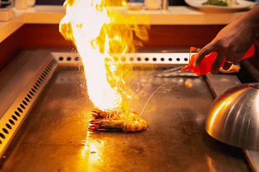 Close-up shot of unrecognizable teppanyaki chef making fire on top of a tiger prawn ,with oil in a flat iron plate, at a Japanese restaurant. The chef making the performance in an open kitchen
