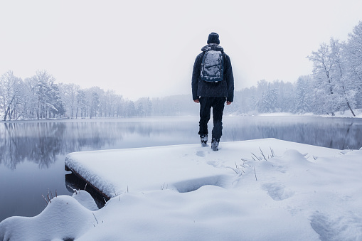 Young man in jacket standing on water gate, pond shore with snow in winter storm. Czech landscape background