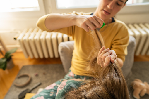 Mother doing head lice cleaning on little daughter while sitting on sofa at home