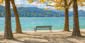 Bench in sunlight at lake Woerthersee under trees in autumn leaf color