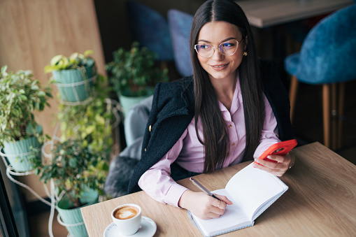 Young businesswoman writing notes while using phone