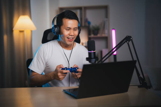 istock Asian influencer man wearing glasses smile cheerful using joystick playing game live streaming greeting video conference with laptop and wearing headphone. Asian man teaching online concept. 1931905698