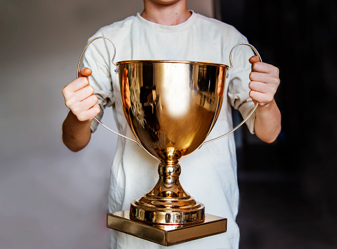 A boy in a white T-shirt holds a golden trophy close-up. Winning success achieve the goal, leadership, award concept