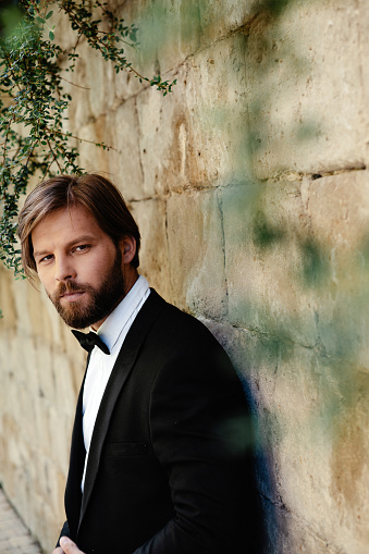 Portrait of caucasian man in trendy black suit posing near wall outdoors. Confident bearded groom looking at camera while standing on fresh air.