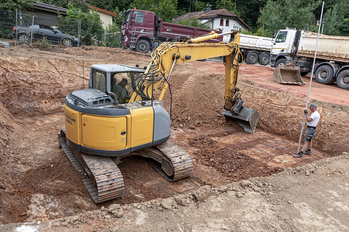 Fischbach, Germany - July 15, 2020: large digger digs a hole for the cellar of a family house,the worker checks the distance and heigh of the excavation pit