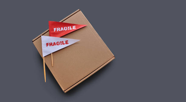 istock Small paper flags with the warning 'Fragile' as a label and empty cardboard box on monochrome gray background 1931944268