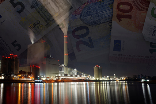 Night shot of a coal-fired power station with reflection in the river and smoking chimneys. A downward gradient of euro banknotes from the night sky as a symbol of rising energy costs.