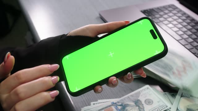 The woman taps the screen and with each tap money appears. View of a woman's hands on a phone with a green screen for copying. Loan, deposit, business and finance concept.