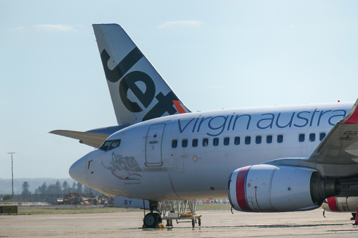 The forward fuselage of a Virgin Australia Boeing B737-7FE plane, registration VH-VBY, parked behind the domestic terminal at Sydney Kingsford-Smith Airport.  In the background is the vertical stabiliser of a Jetstar Airbus A320-232 plane, registration VH-XNP.  This image was taken from Ross Smith Avenue, Mascot, on a hot and sunny afternoon on 13 January 2024.